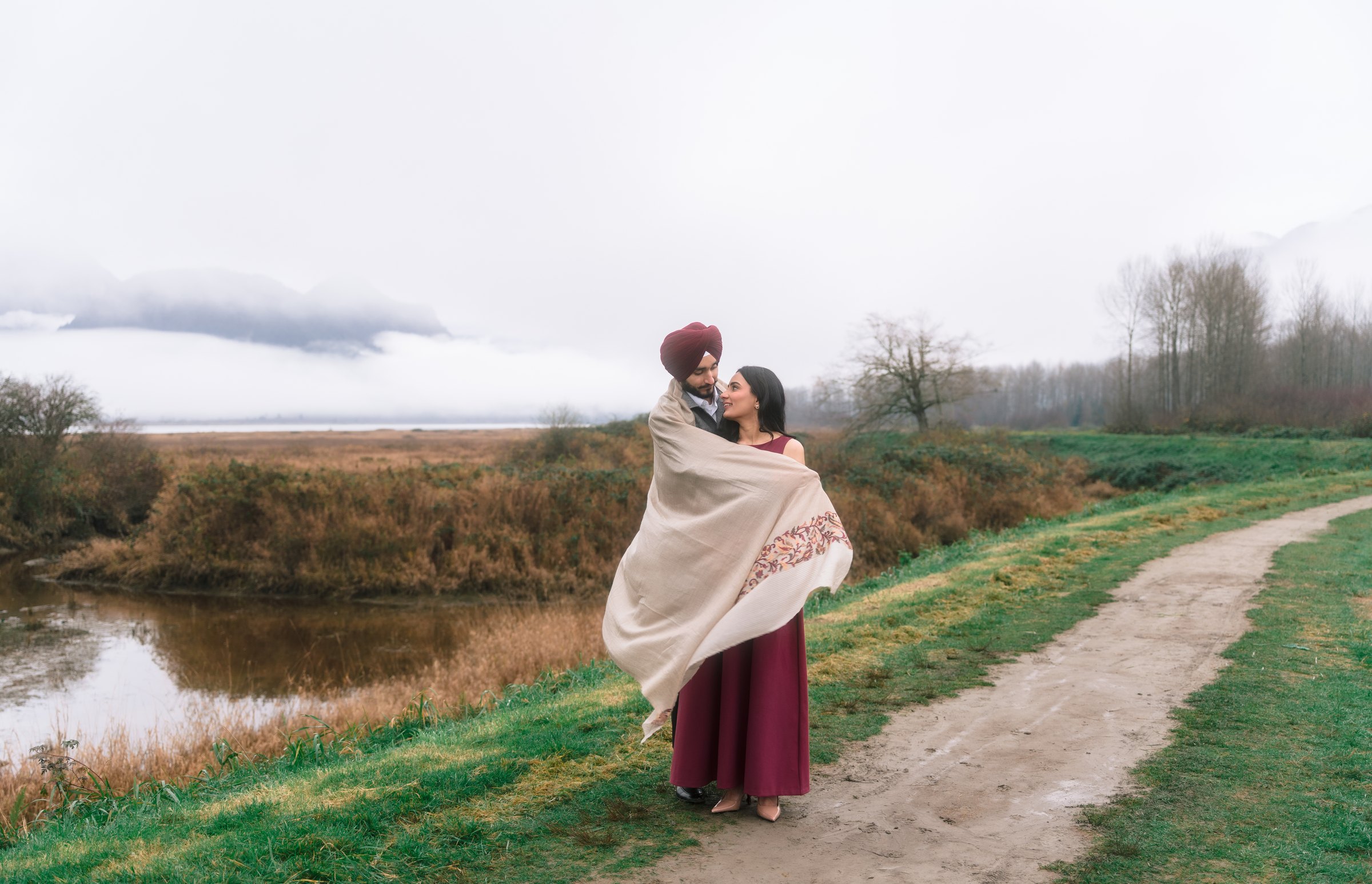 Simar & Manjeet wrapped in an embroidered cashmere shawl overlooking tidal waters and mountain reflections at Pitt Lake in Pitt Meadows, BC — White Daisy Weddings